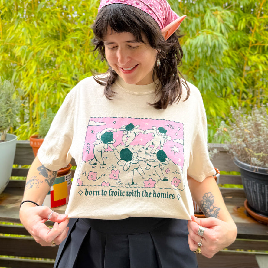 Person wearing a t-shirt with a graphic design and text, standing outdoors with plants in the background.